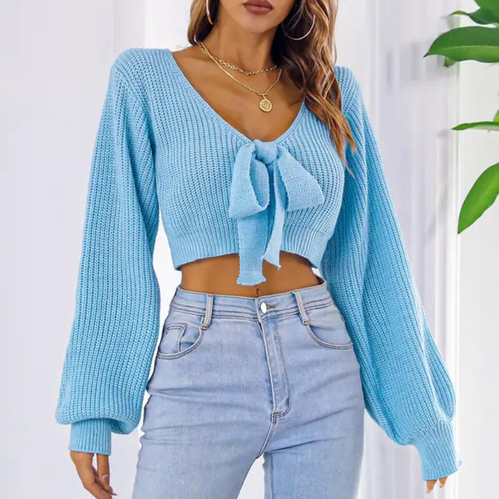 Vrouw draagt lichtblauwe gebreide trui met V-hals en strik, gecombineerd met high-waisted jeans. Trendy damesmode, casual outfit, herfstkleding.