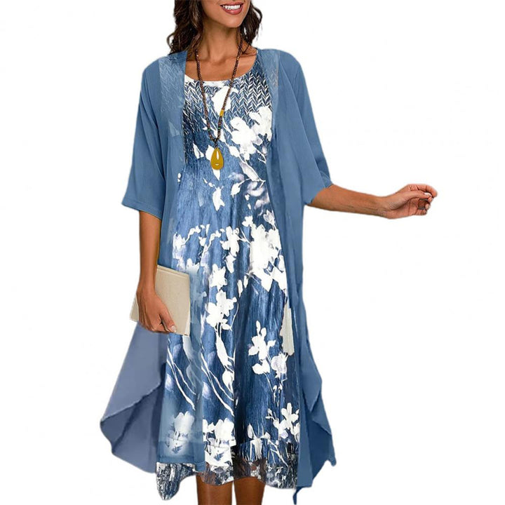Vrouw in blauwe zomerjurk met bloemenprint en driekwart mouwen, draagt een ketting en houdt een clutch. Perfecte outfit voor casual zomerdagen.