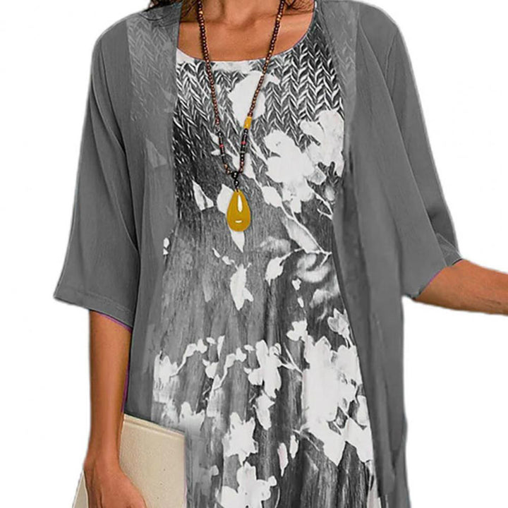 Grijze zomerjurk met bloemenprint en driekwart mouwen, gecombineerd met een lange ketting en een beige clutch. Perfect voor casual damesmode.
