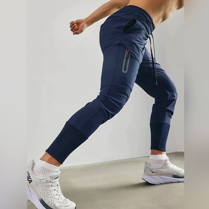 Man in sportieve joggingbroek voor heren, donkerblauw, met ritszakken en elastische tailleband, draagt witte sportschoenen, fitnesskleding.