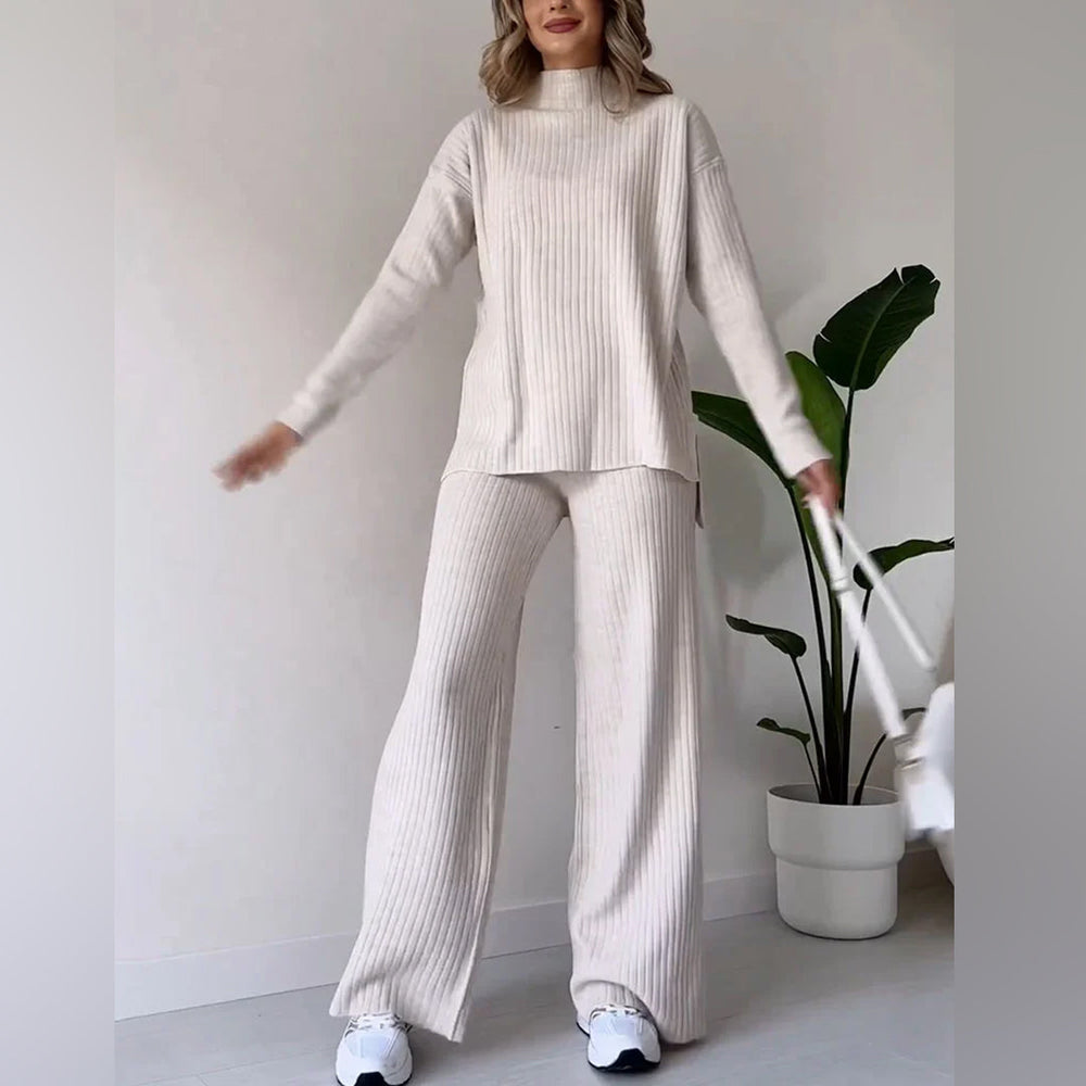 Vrouw in beige ribgebreide loungewear set, bestaande uit trui en wijde broek, met witte sneakers en handtas, naast groene kamerplant.