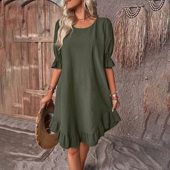 Vrouw in een olijfgroene zomerjurk met ruches, staand voor een rustieke houten deur. Perfecte boho-chic outfit voor casual zomerdagen.