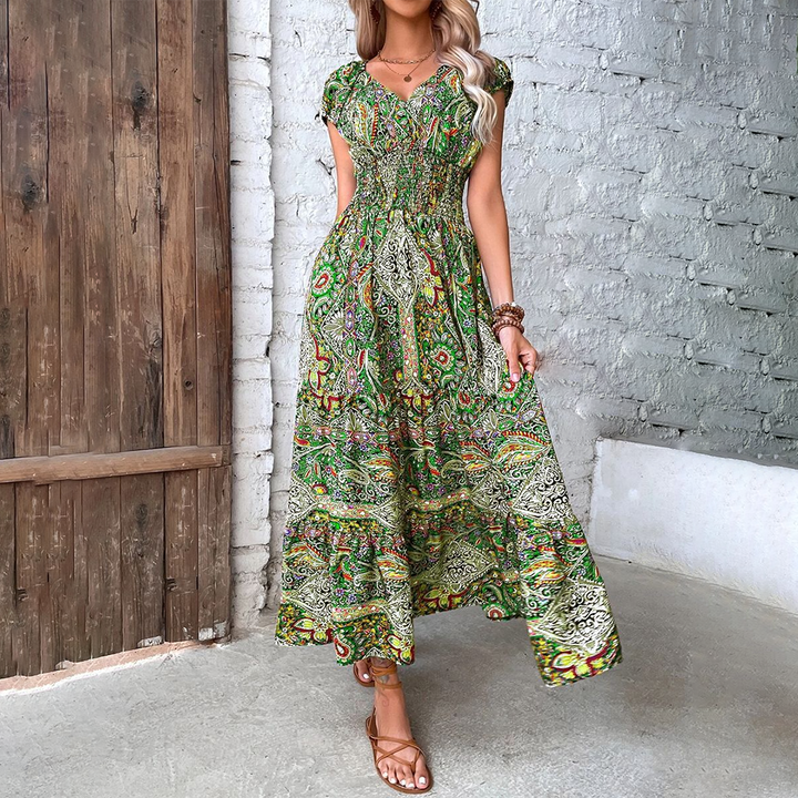 Vrouw in lange, groene bohemian jurk met kleurrijk paisley-patroon, staand voor houten deur en witte bakstenen muur. Zomermode, boho-stijl.
