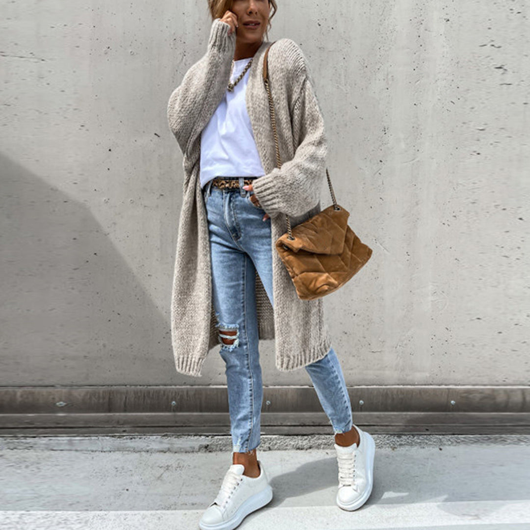 Vrouw in casual outfit met beige gebreid vest, witte sneakers en blauwe gescheurde jeans, poseert tegen een betonnen muur. Modieuze herfstkleding.
