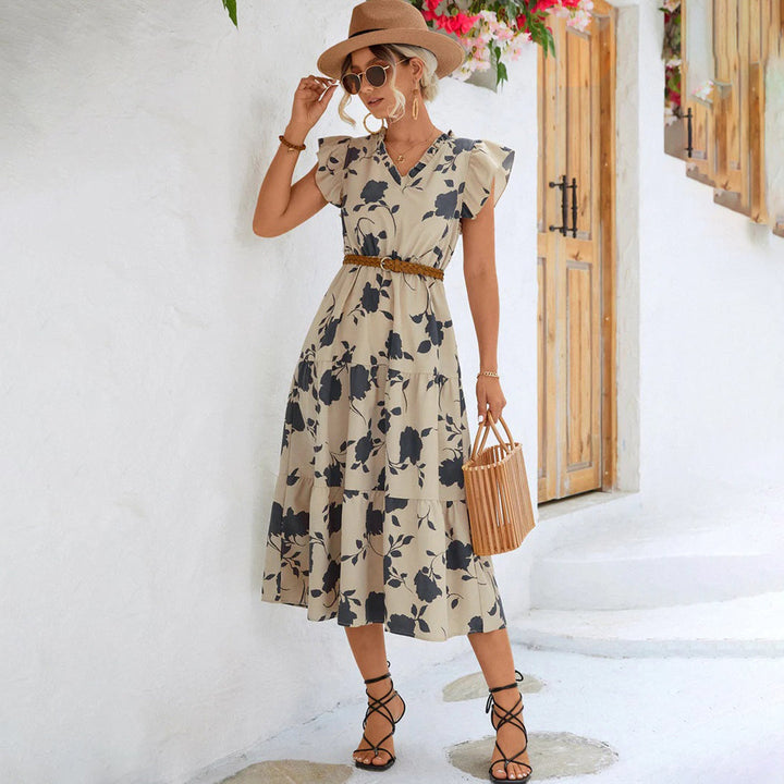 Vrouw in beige zomerjurk met bloemenprint, rieten tas en hoed, poseert buiten. Perfecte boho-chic outfit voor warme dagen.