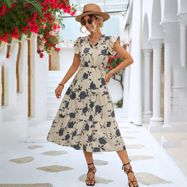Vrouw in beige zomerjurk met bloemenprint, zonnehoed en sandalen, poseert in mediterrane straat. Zomerjurk dames, casual mode, vakantie outfit.