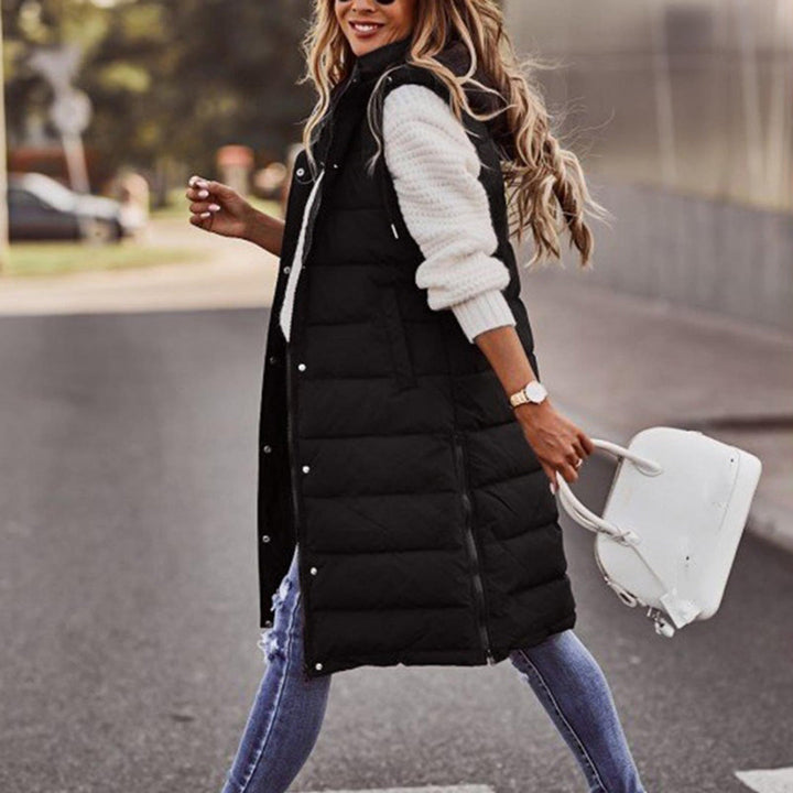 Vrouw in zwarte gewatteerde lange bodywarmer, witte trui en blauwe jeans, draagt witte handtas. Modieuze herfstkleding voor dames.