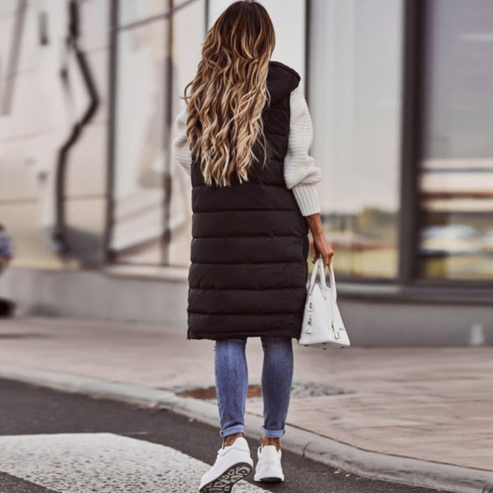 Vrouw in zwarte gewatteerde lange bodywarmer, blauwe jeans en witte sneakers, draagt witte handtas. Stijlvolle herfstmode op straat.