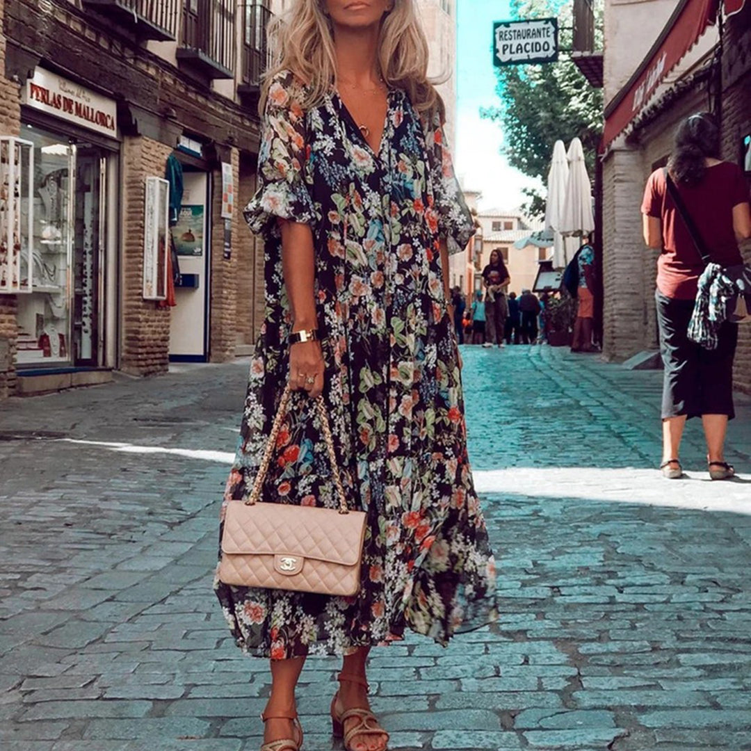 Vrouw in kleurrijke bloemenjurk met beige handtas, wandelend door pittoreske straat. Zomermode, bohemian stijl, stedelijke achtergrond.