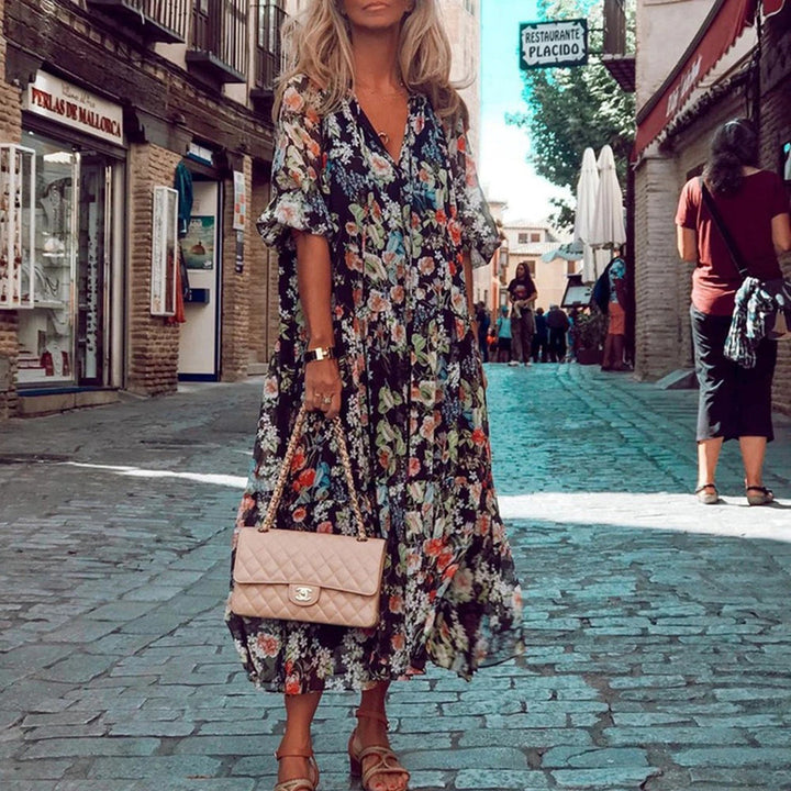 Vrouw in kleurrijke bloemenjurk met beige handtas, wandelend door pittoreske straat. Zomermode, bohemian stijl, stedelijke achtergrond.