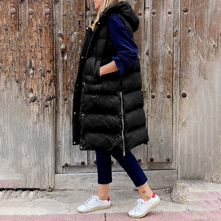 Vrouw in zwarte gewatteerde lange bodywarmer met capuchon, blauwe broek en witte sneakers, staand voor een houten deur. Wintermode, casual outfit.