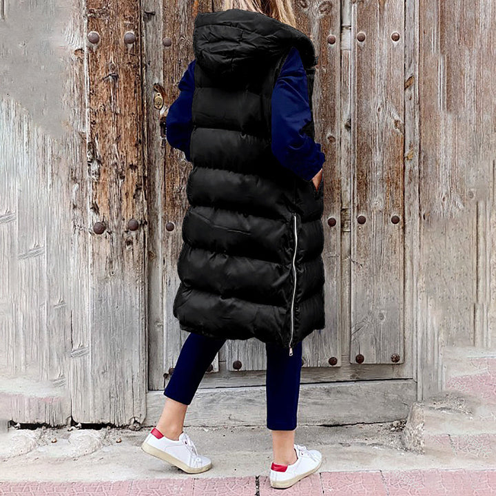 Vrouw in zwarte gewatteerde lange bodywarmer met capuchon, blauwe broek en witte sneakers, staand voor een rustieke houten deur. Wintermode, casual outfit.