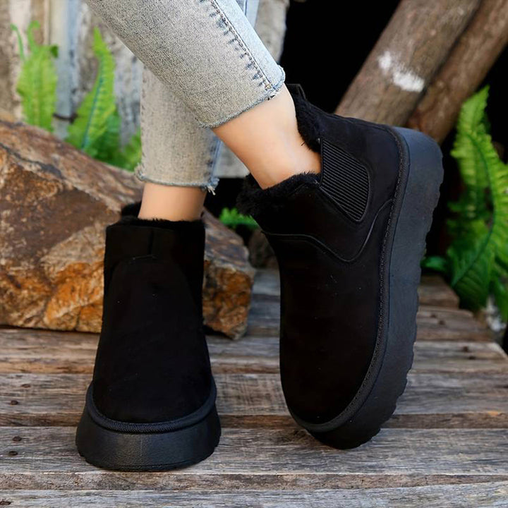 Zwarte suède enkellaarsjes met bontvoering, elastische zijkanten en dikke zool, gedragen door model op houten achtergrond. Wintermode, comfortabele schoenen.