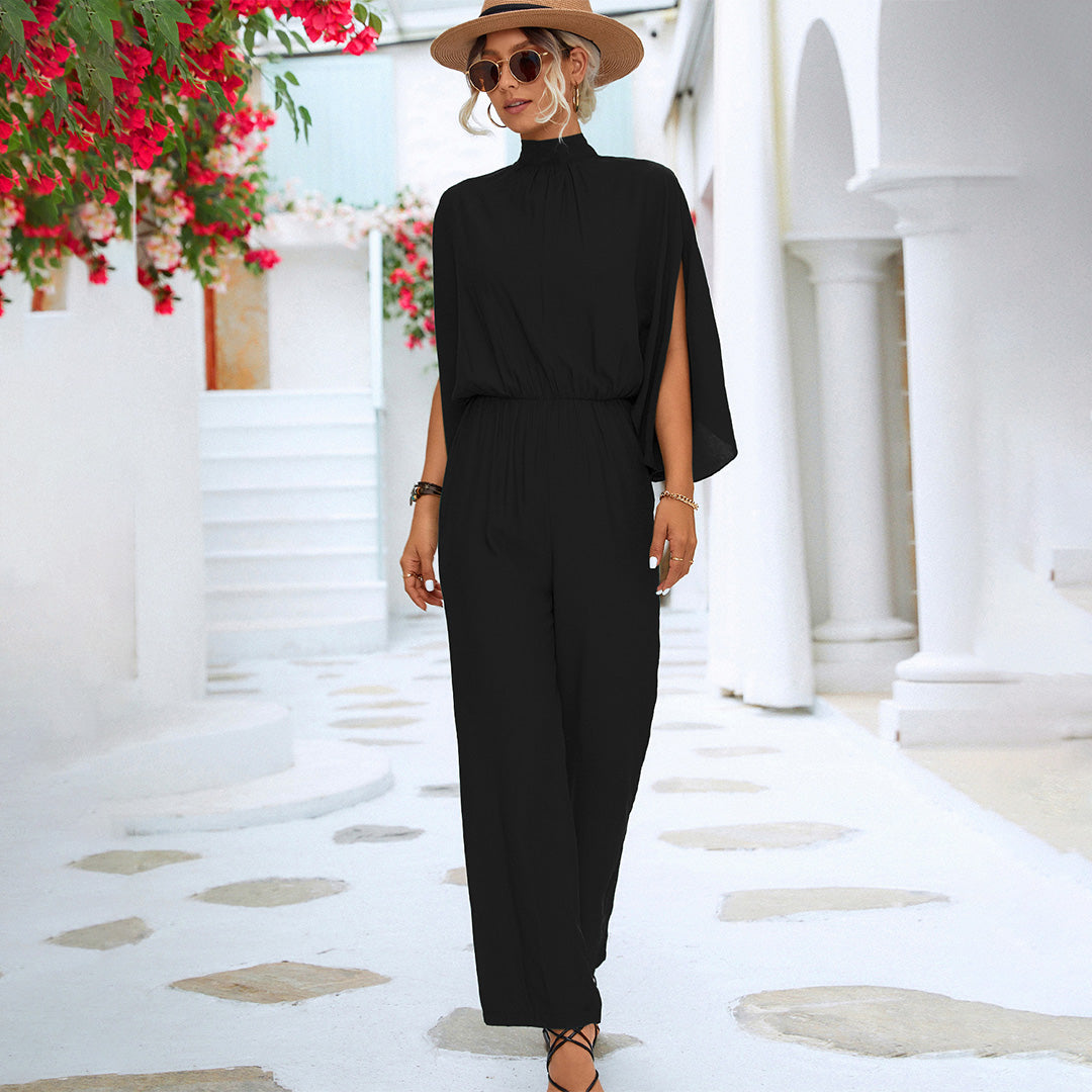 Vrouw in elegante zwarte jumpsuit met hoge hals en cape-mouwen, draagt zonnebril en hoed, poseert in mediterrane straat met witte architectuur.