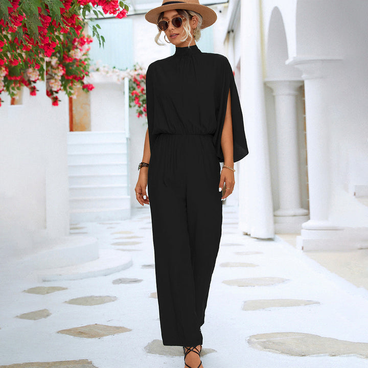 Vrouw in elegante zwarte jumpsuit met hoge hals en cape-mouwen, draagt zonnebril en hoed, poseert in mediterrane straat met witte architectuur.