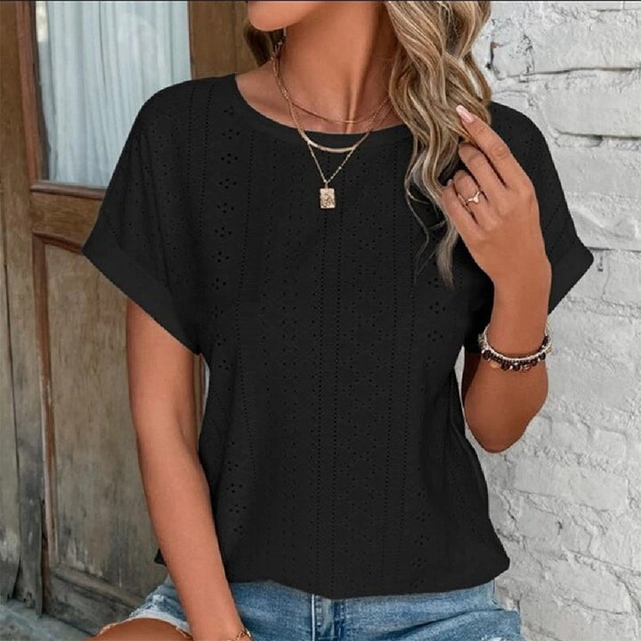Vrouw draagt zwarte kanten blouse met korte mouwen, gouden ketting en armband, casual zomeroutfit, tegen een witte bakstenen muur.