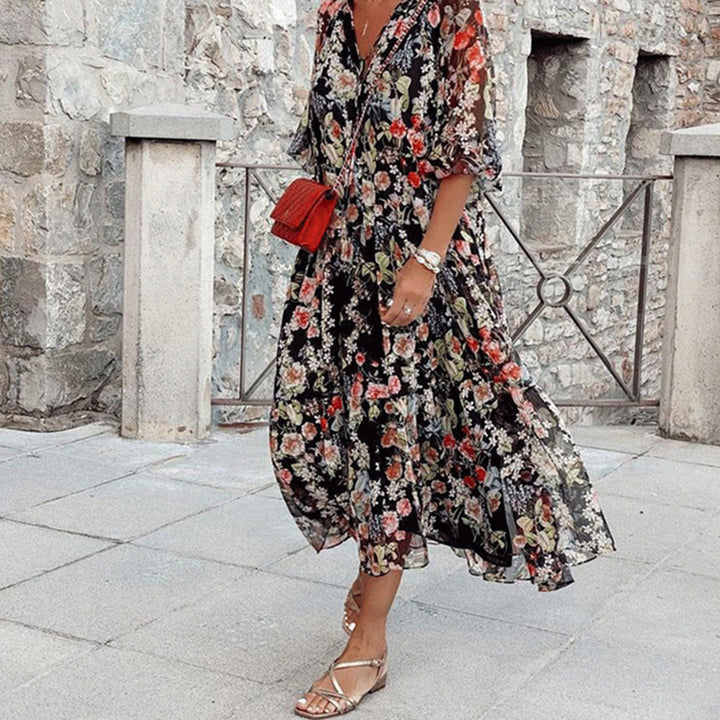 Vrouw in lange, zwarte bloemenjurk met rode tas en sandalen, staand voor stenen muur. Bohemian stijl, zomerjurk, casual mode.