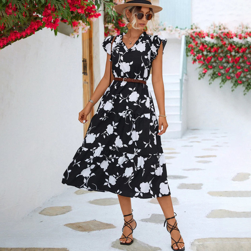 Vrouw in zwarte bloemenjurk met hoed, wandelend op geplaveide straat met bloemen. Zomerse mode, elegante dameskleding, casual chique stijl.