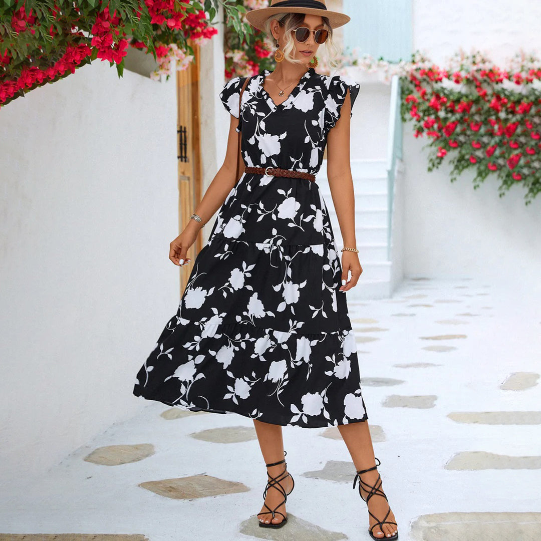 Vrouw in zwarte bloemenjurk met hoed, wandelend op geplaveide straat met bloemen. Zomerse mode, elegante dameskleding, casual chique stijl.