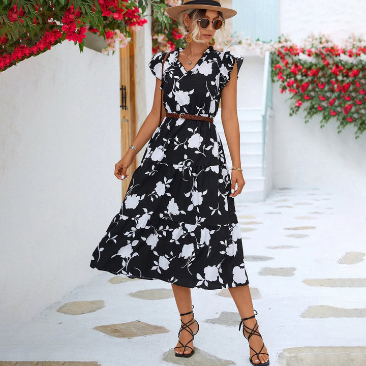 Vrouw in zwarte bloemenjurk met hoed, wandelend op geplaveide straat met bloemen. Zomerse mode, elegante dameskleding, casual chique stijl.