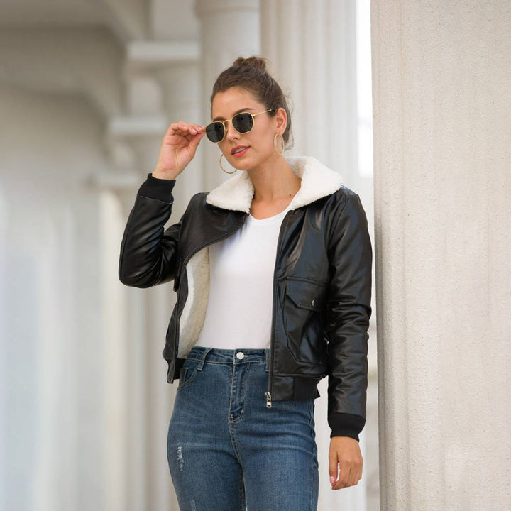 Vrouw in zwarte leren jas met witte kraag, spijkerbroek en zonnebril leunt tegen zuil. Modieuze dameskleding, herfststijl, casual outfit.