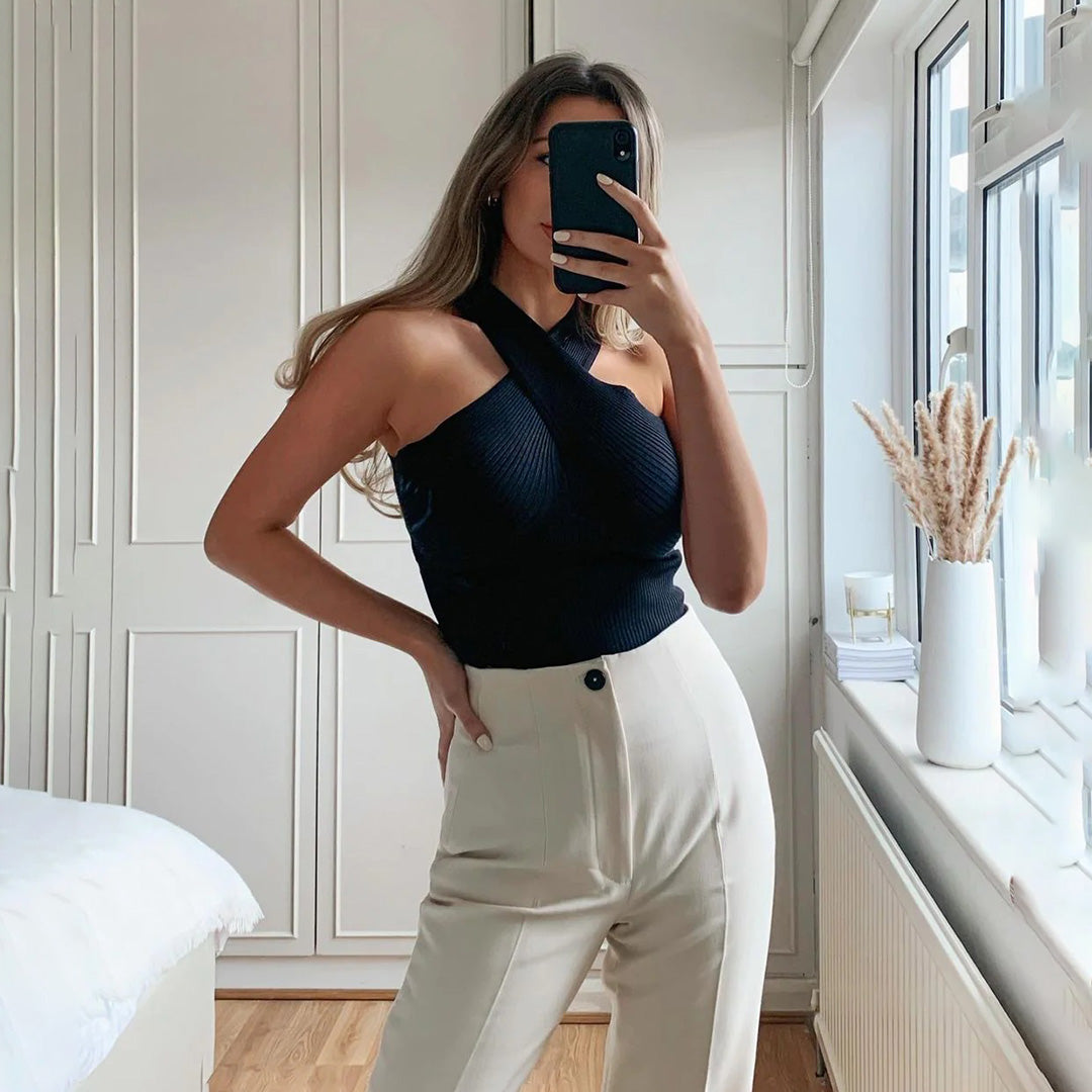 Vrouw in moderne slaapkamer draagt zwarte asymmetrische top en beige hoge taille broek, poseert voor spiegel. Stijlvolle mode, minimalistisch interieur.