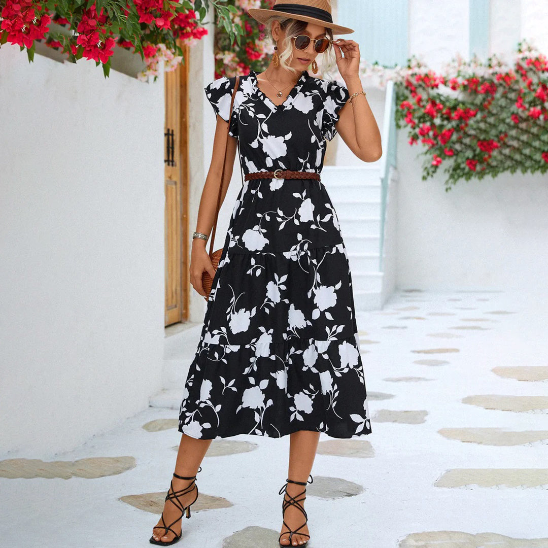 Vrouw in zwarte bloemenjurk met hoed poseert buiten, omgeven door rode bloemen en witte muren. Zomerse mode, elegante dameskleding, casual stijl.