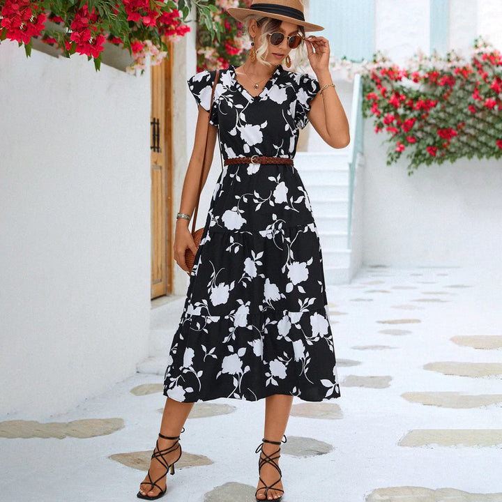 Vrouw in zwarte bloemenjurk met hoed poseert buiten, omgeven door rode bloemen en witte muren. Zomerse mode, elegante dameskleding, casual stijl.