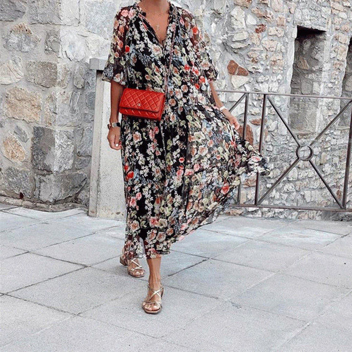 Vrouw in lange, zwarte bloemenjurk met rode schoudertas en sandalen, wandelend op stenen pad voor stenen muur. Zomermode, casual elegantie.