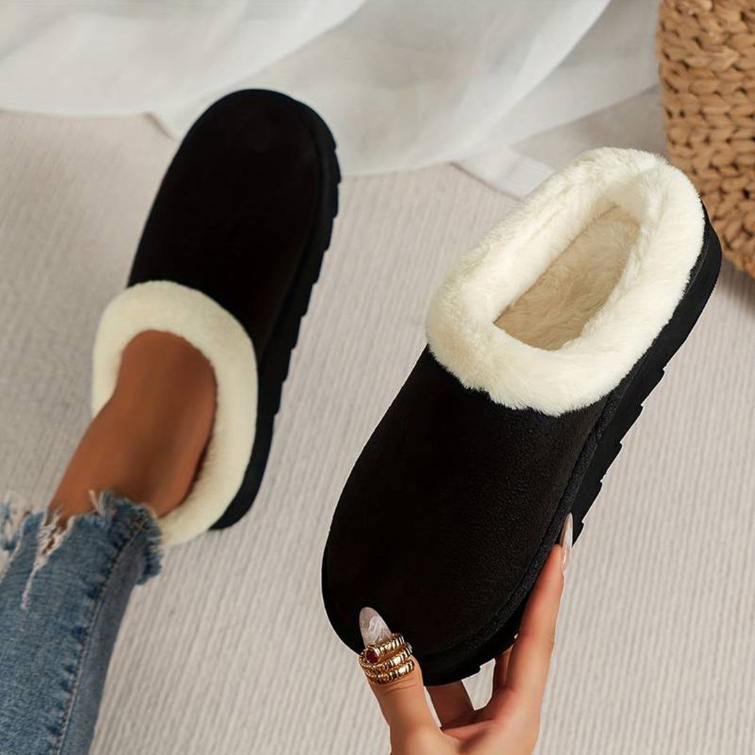 Zwarte pantoffels met witte voering, gedragen door persoon met spijkerbroek. Comfortabele winterse huisschoenen, ideaal voor warme voeten thuis.