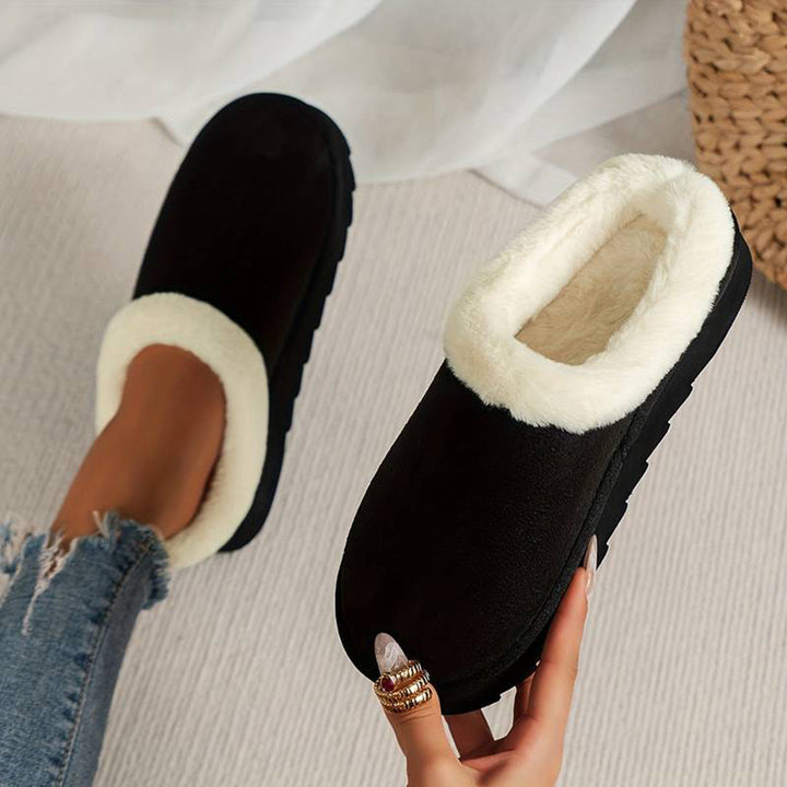 Zwarte pantoffels met witte voering, gedragen door persoon met spijkerbroek. Comfortabele winterse huisschoenen, ideaal voor warme voeten thuis.