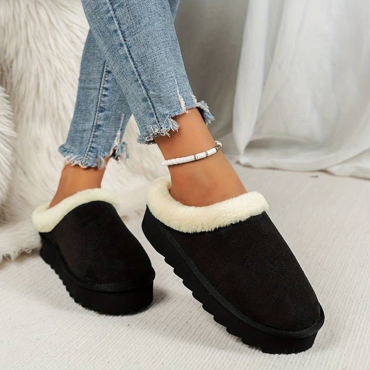 Zwarte dames pantoffels met witte voering, gedragen met gescheurde jeans. Comfortabele winterse huisschoenen, ideaal voor warme voeten thuis.