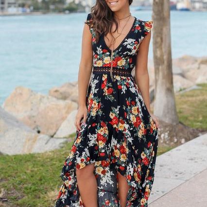 Vrouw in zwarte bloemenjurk met rode en witte bloemen, staand bij waterkant. Zomerse boho-stijl, casual mode, strandoutfit.