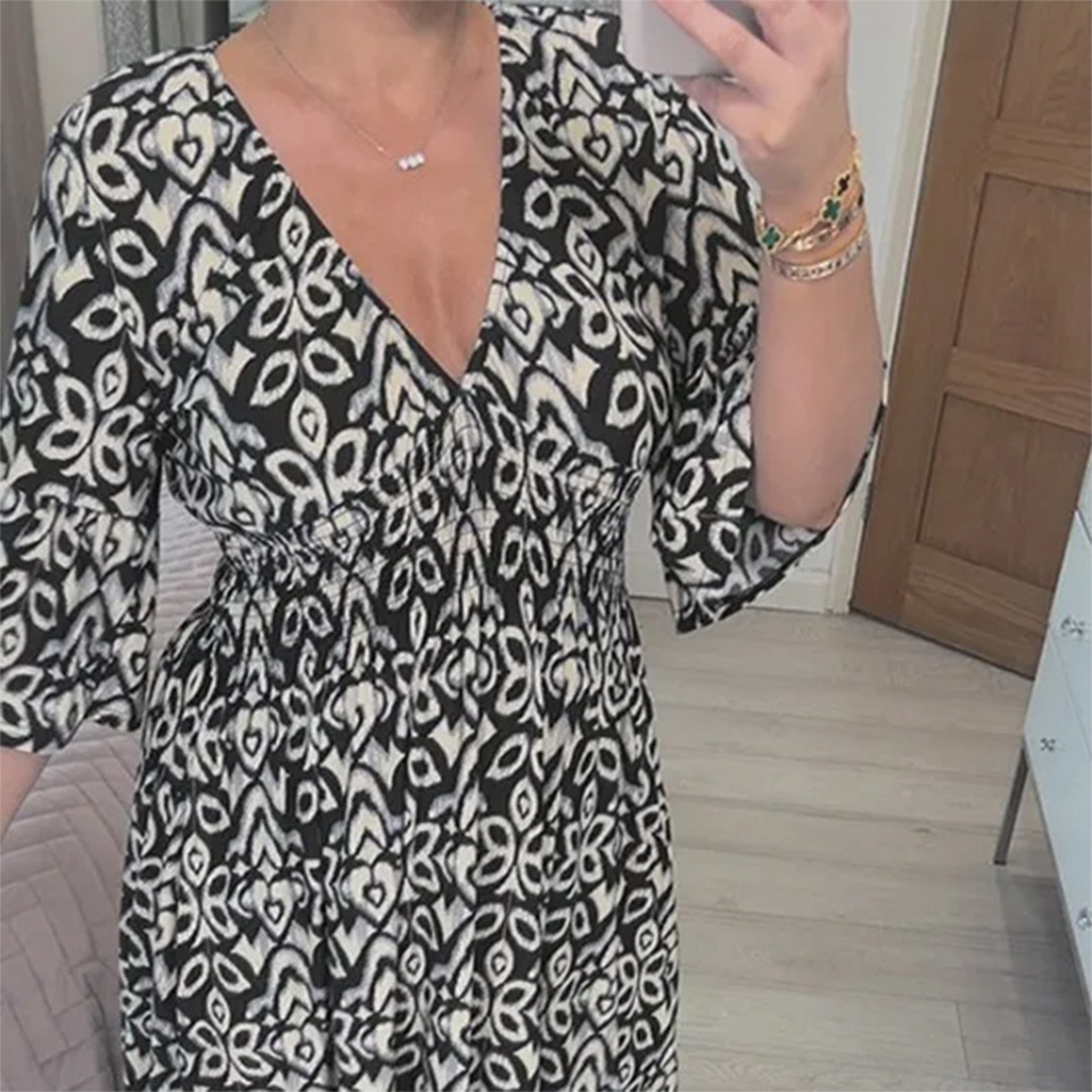 Vrouw in zwart-witte jurk met geometrisch patroon, V-hals en driekwartsmouwen, maakt selfie in moderne slaapkamer. Trendy damesmode, casual stijl.