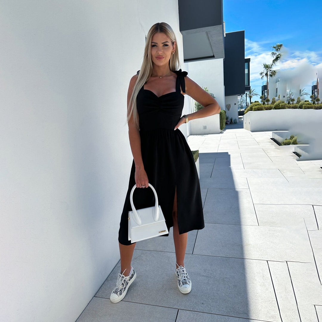 Vrouw in zwarte zomerjurk met strikbandjes, witte sneakers en witte handtas, staand op moderne buitenlocatie. Zomerjurk, mode, dameskleding.