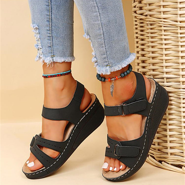 Zwarte damesplatformsandalen met verstelbare bandjes, gedragen met gerafelde jeans en kleurrijke enkelbandjes, naast een gevlochten mand.