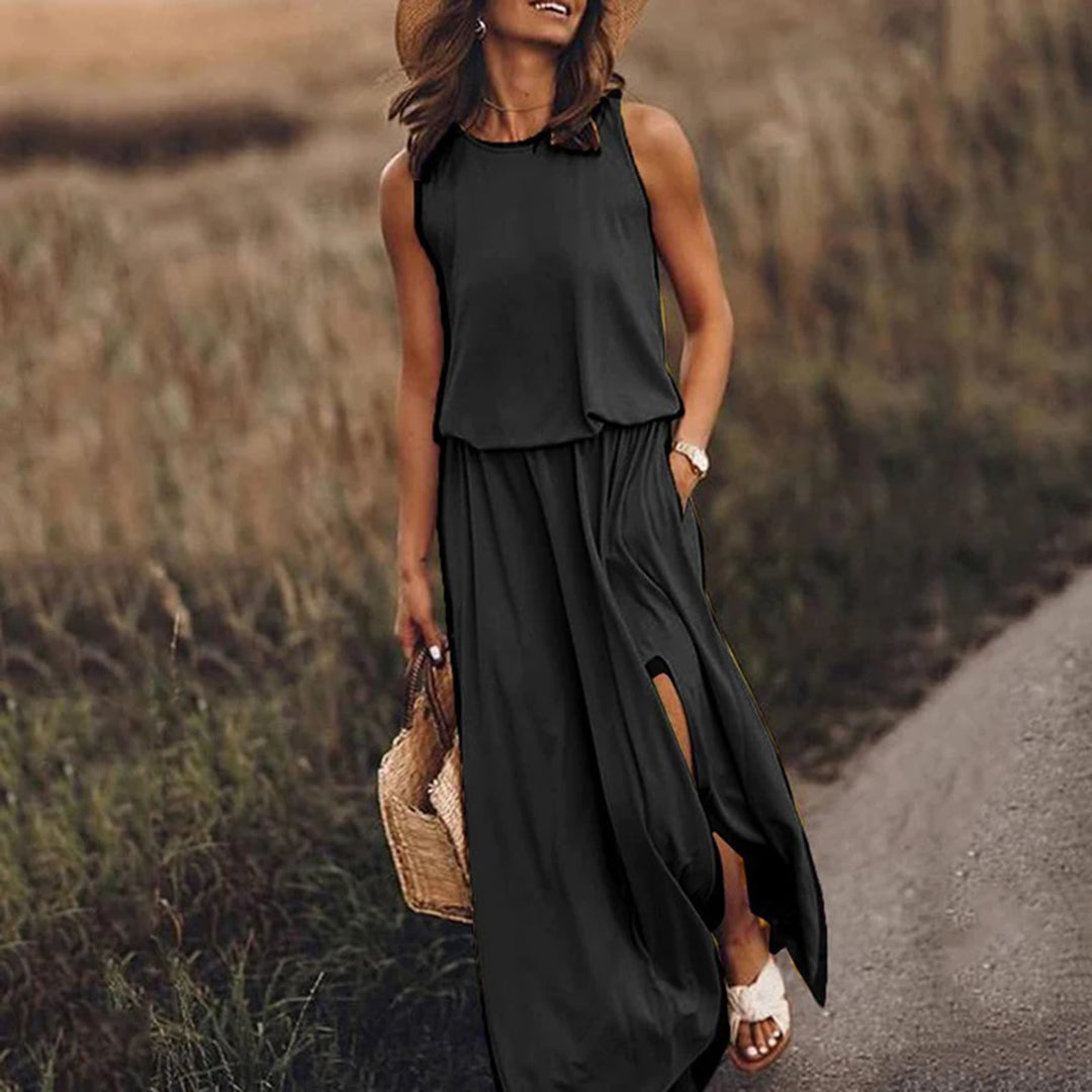 Vrouw in zwarte maxi-jurk met hoge split, strohoed en rieten tas, wandelend op landweg. Zomeroutfit, casual mode, dameskleding.