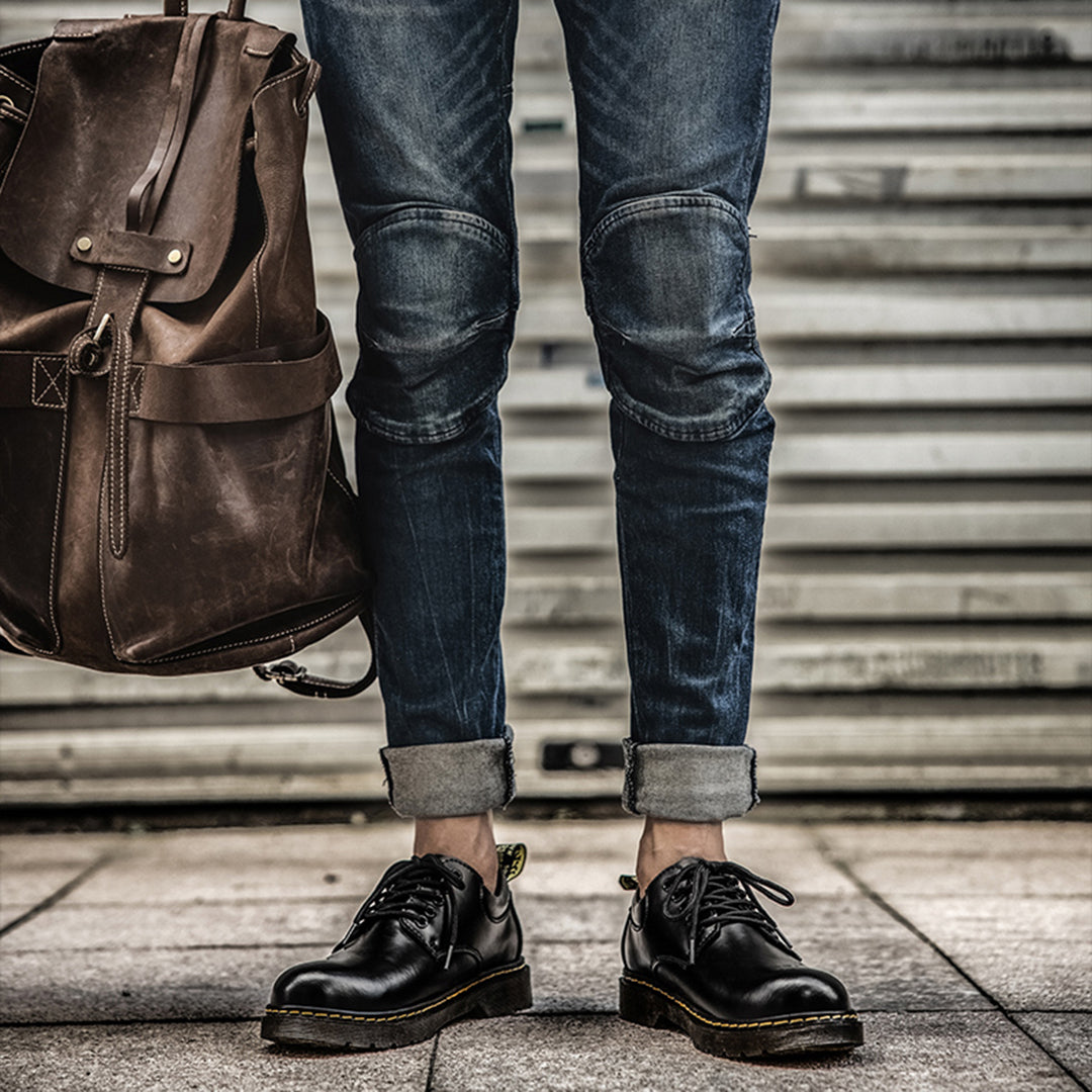 Zwarte leren schoenen met veters, gedragen door persoon in opgerolde jeans, naast bruine leren rugzak. Stijlvolle herenschoenen, casual mode.