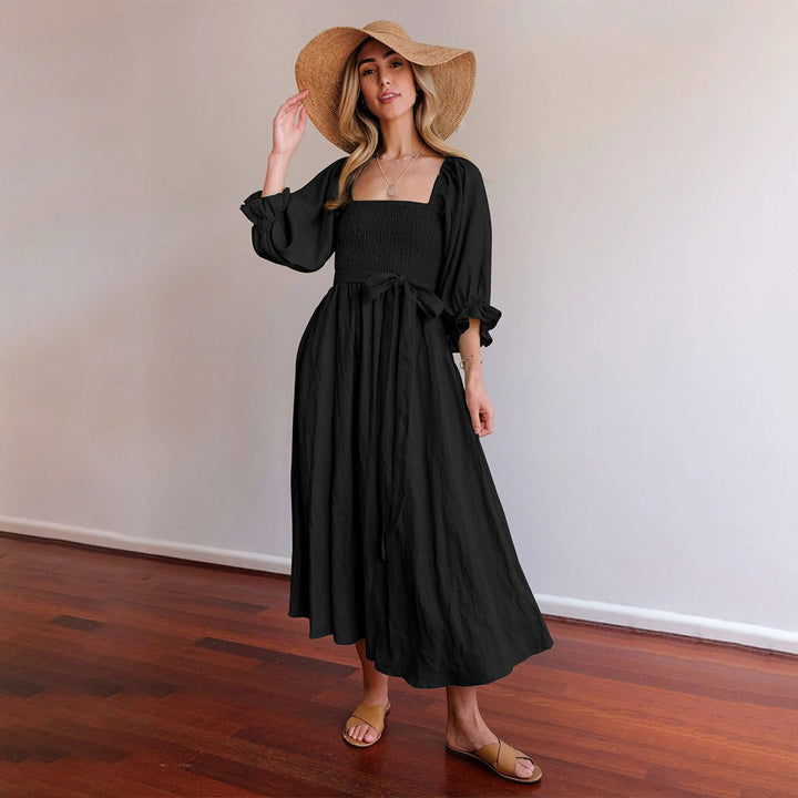 Vrouw in zwarte maxi-jurk met strohoed, staand op houten vloer. Elegante damesmode, zomeroutfit, casual chique stijl.