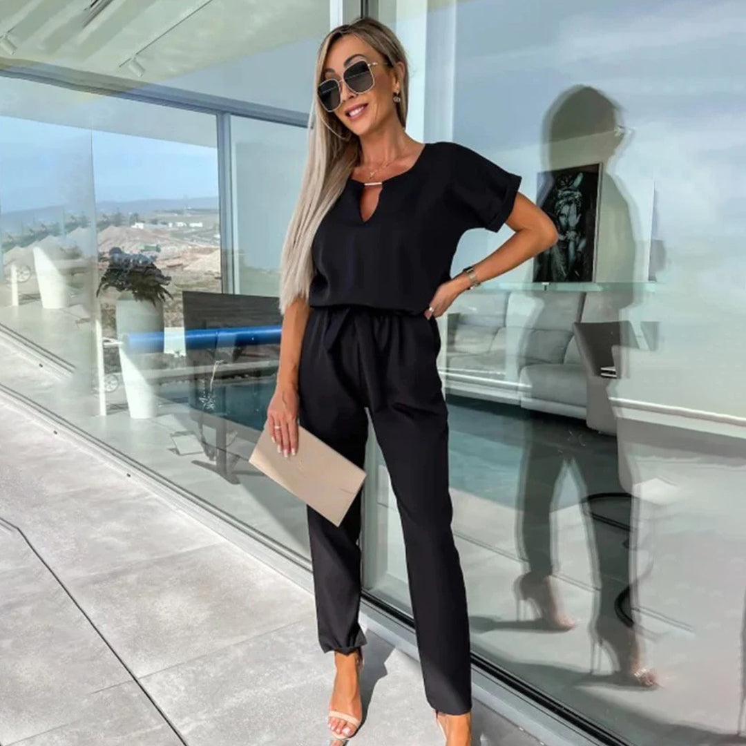 Vrouw in stijlvolle zwarte jumpsuit met zonnebril en beige clutch, staand op modern terras met glazen wand. Perfecte zomeroutfit voor dames.