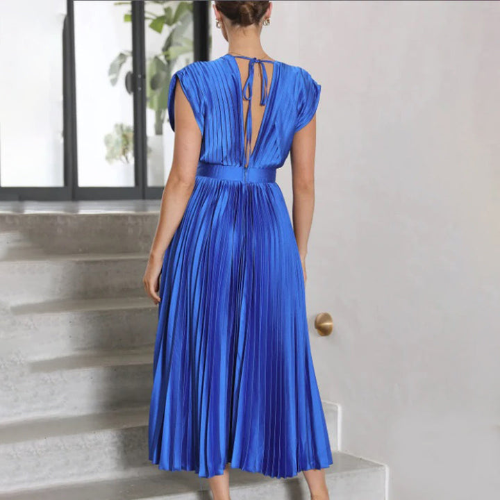 Vrouw in elegante blauwe geplooide jurk, rugaanzicht, staand op trap bij raam. Perfect voor avondkleding, modetrends, stijlvolle damesmode.