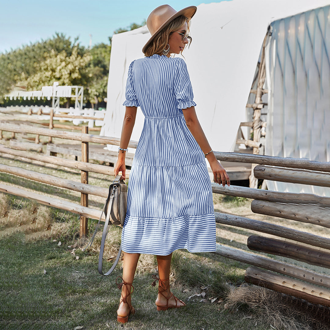 Vrouw in blauwe gestreepte zomerjurk en hoed, staand bij houten hek op zonnige dag. Mode, zomerjurk, buiten, casual stijl.