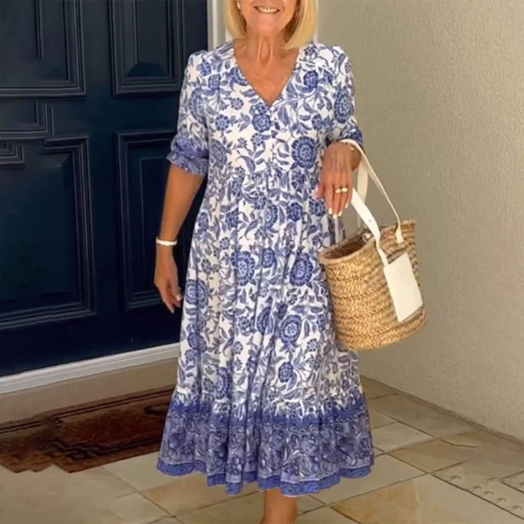 Vrouw in blauwe bloemenjurk met rieten tas, staand voor een donkerblauwe deur. Zomerse outfit, casual mode, dameskleding.