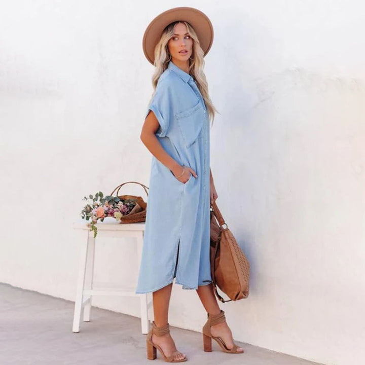 Vrouw in lichtblauwe denim jurk met korte mouwen, bruine hoed en sandalen, poseert naast tafel met bloemen en bruine tas. Zomerse mode, casual stijl.
