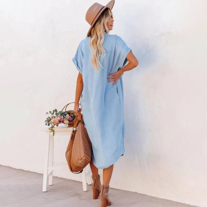 Vrouw in lichtblauwe zomerjurk met hoed en bruine tas, staand voor witte muur. Casual damesmode, boho-stijl, zomerkleding.