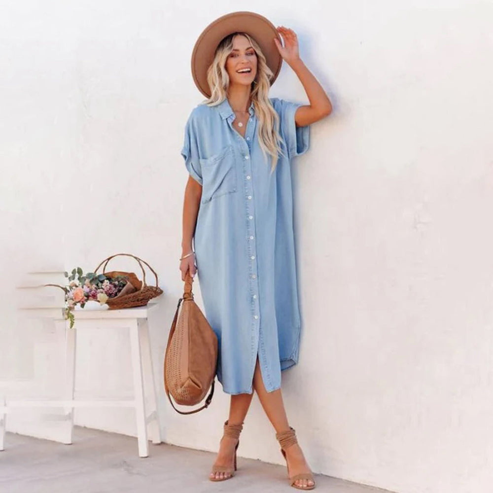 Vrouw in lichtblauwe denim jurk met knopen, draagt een bruine hoed en tas, poseert tegen witte muur. Zomerse mode, casual stijl.