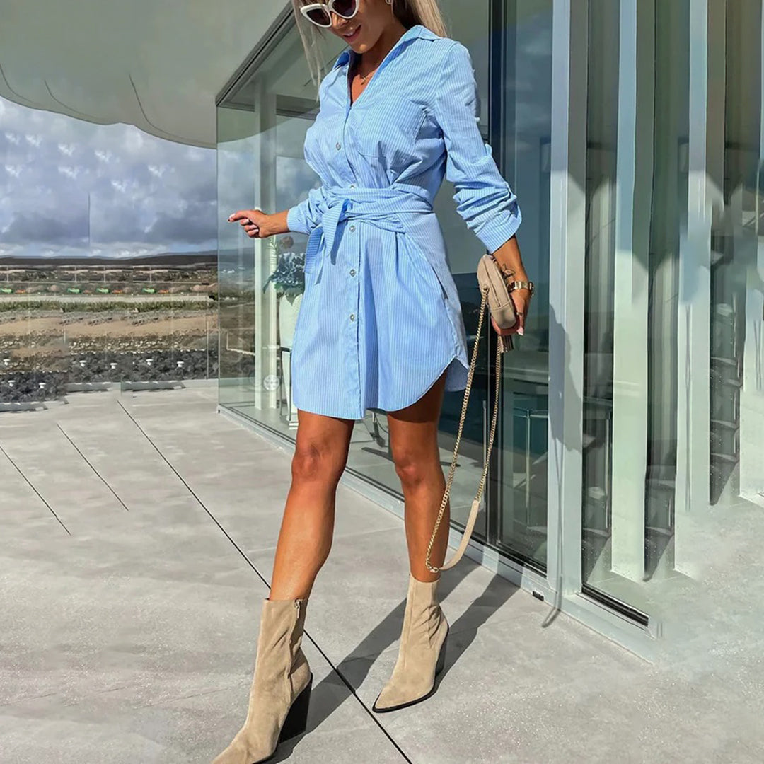Vrouw in blauwe overhemdjurk met ceintuur, beige laarzen en zonnebril, poseert buiten bij glazen gebouw. Zomermode, casual chic stijl.