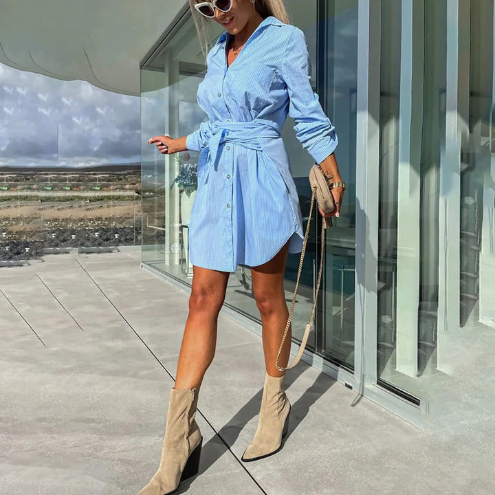Vrouw in blauwe overhemdjurk met ceintuur, beige laarzen en zonnebril, poseert buiten bij glazen gebouw. Zomermode, casual chic stijl.