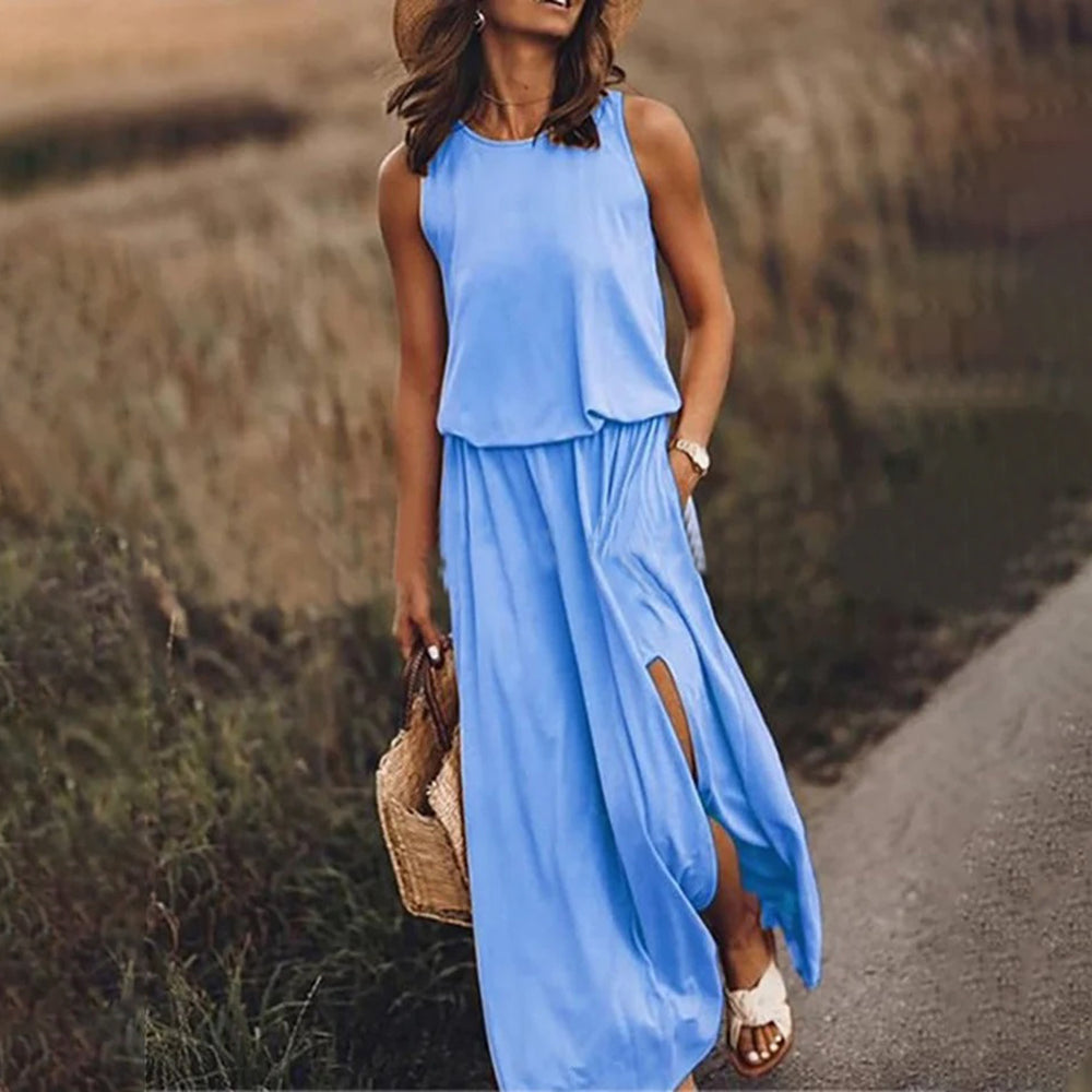 Vrouw in blauwe zomerjurk met hoge split, strohoed en rieten tas, wandelend op landweg. Perfecte outfit voor zomerse dagen en strandvakanties.