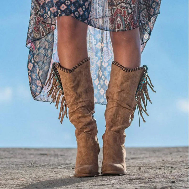 Bruine suède laarzen met franjes, gedragen door een vrouw in een boho-jurk. Perfect voor herfstmode en bohemian stijl. Outdoor achtergrond.
